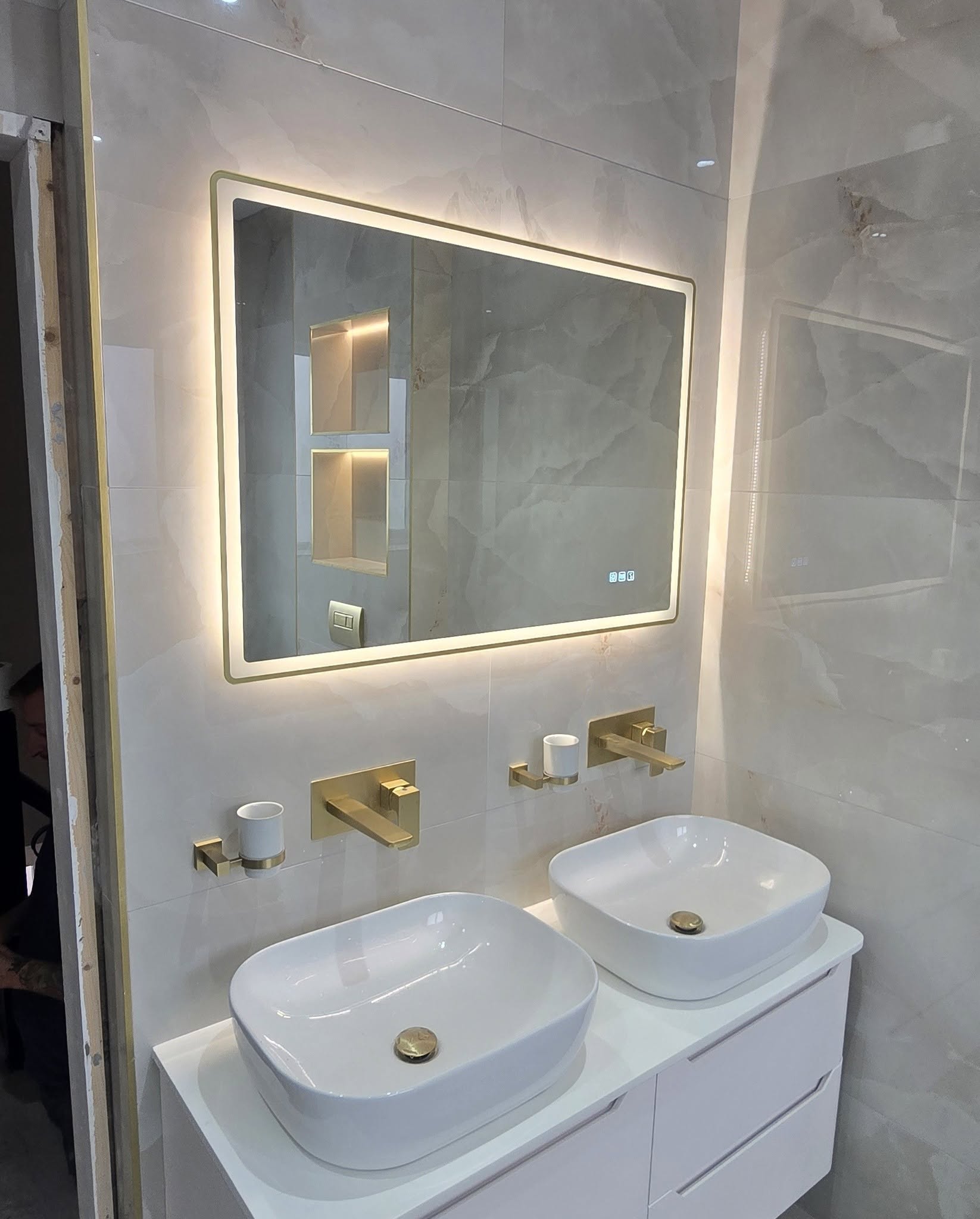Double vessel basins with brushed-brass taps beneath an illuminated mirror and a lit wall niche