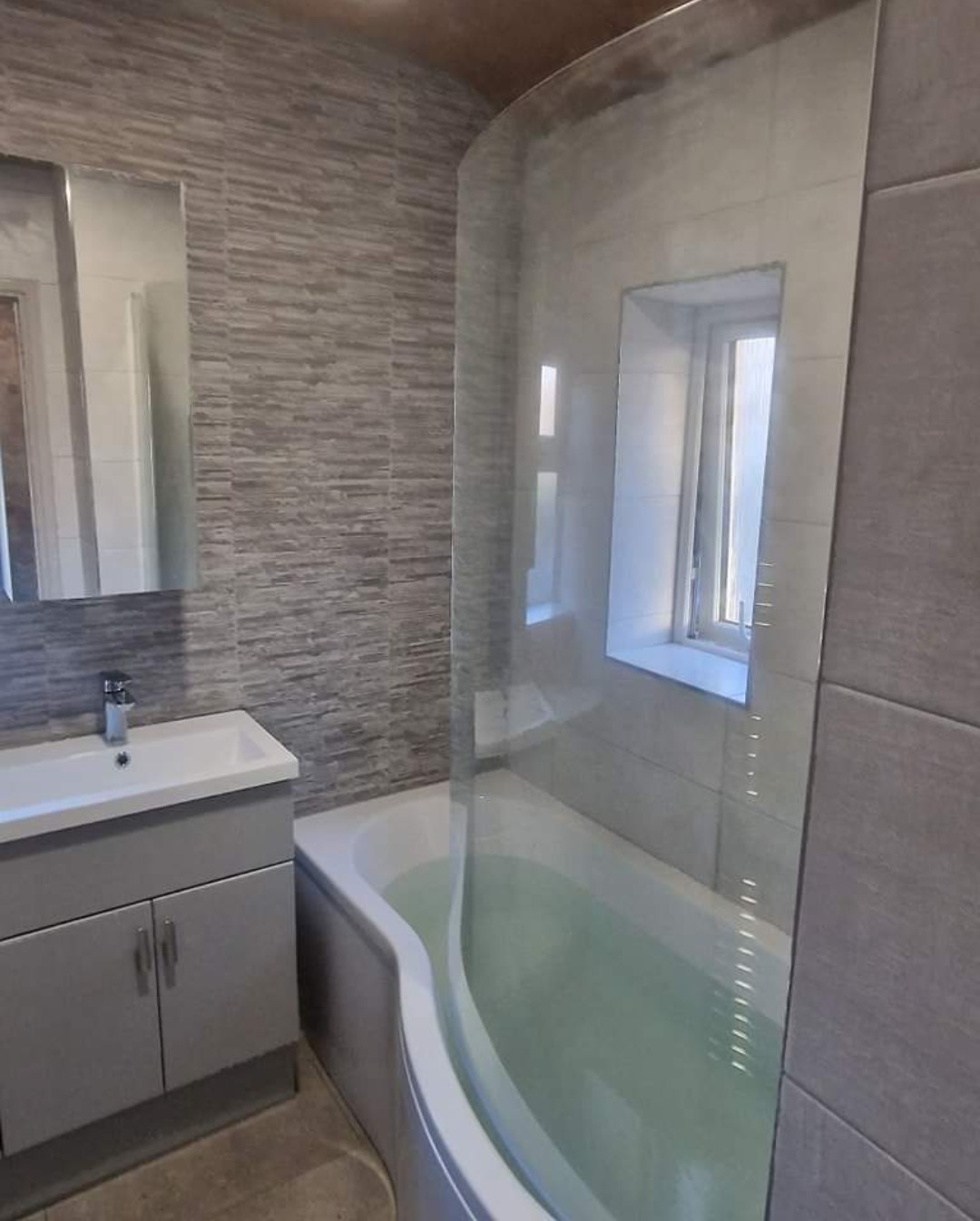 Grey vanity unit beside a bath with a curved shower screen and feature tile wall